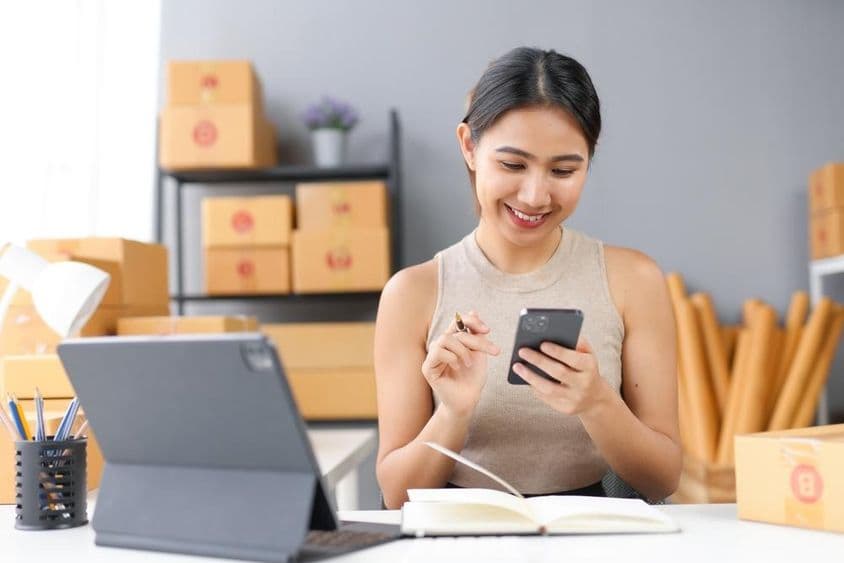 Mosolygó nő okostelefont használ a munkaterületén, körülötte kellékes dobozokkal.